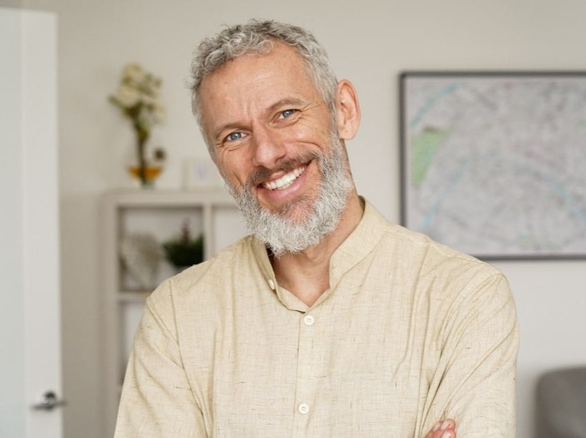 An older man with a beard wearing an off-white button-up shirt smiling